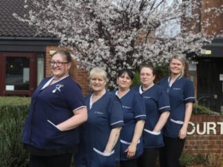 Pictured is of five caring care workers outside, stood together and smiling towards the camera in their navy uniforms.